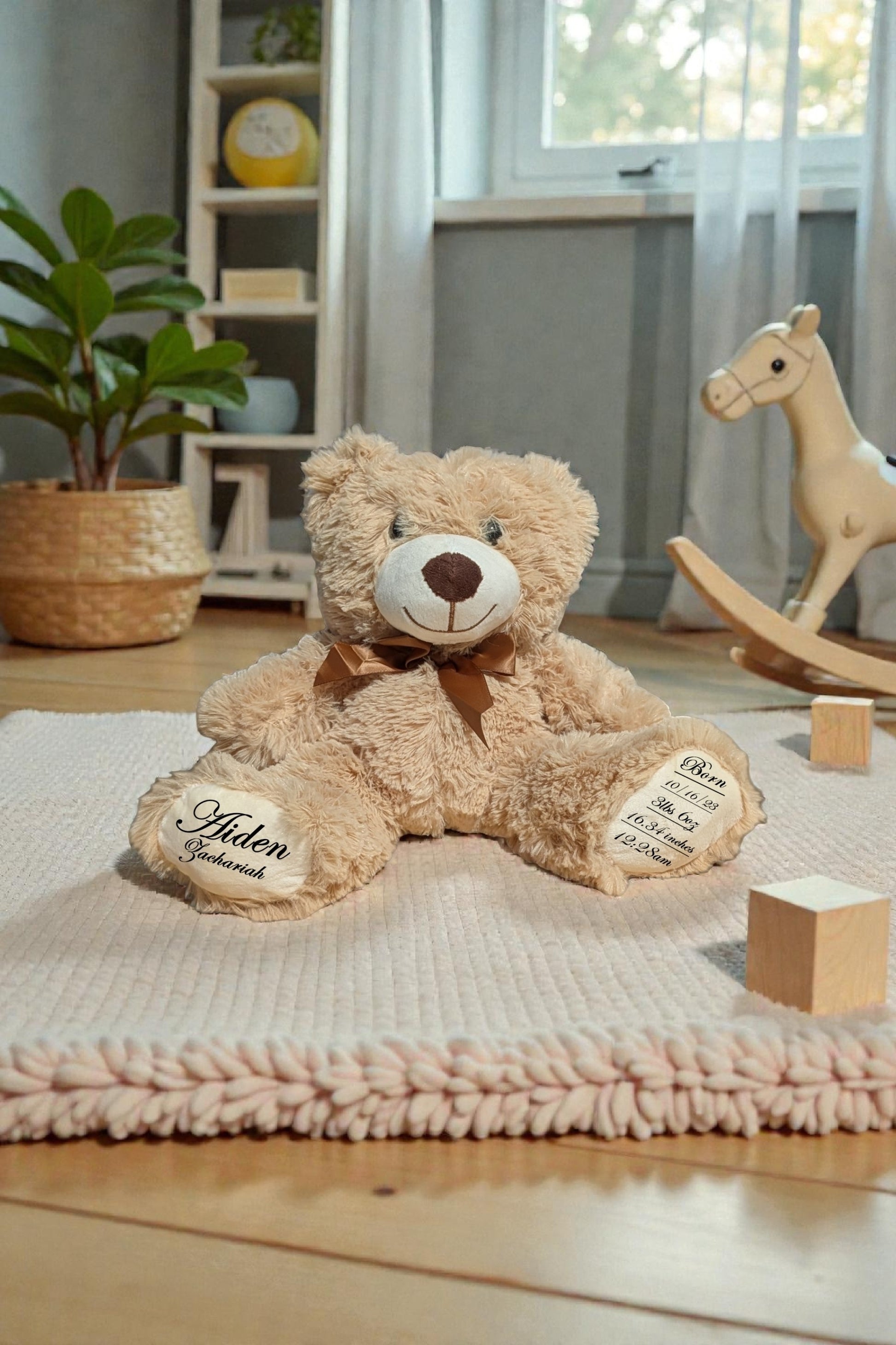Teddy bear with a bow tie sitting on a blanket in a room with toys and plants. The Bears bottoms of the feet have personalized birth stats on them.