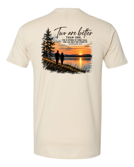 A cream-colored short-sleeve t-shirt shown from the back, featuring a large graphic print of a couple walking at sunset next to a pine tree, with the text 'Two are better than one.'.