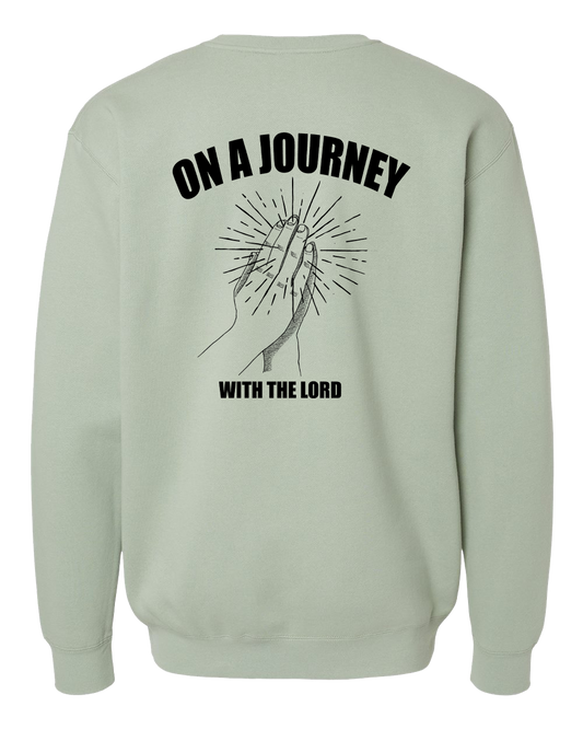 A sage green crewneck sweatshirt shown from the back, featuring a black graphic of praying hands with radiating lines and the text 'ON A JOURNEY WITH THE LORD'.