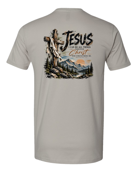 A gray crewneck t-shirt shown from the back, featuring a large graphic print of a wooden cross with a draped white cloth, mountains, pine trees, and the text 'JESUS I CAN DO ALL THINGS THROUGH Christ WHO STRENGTHENS ME'.